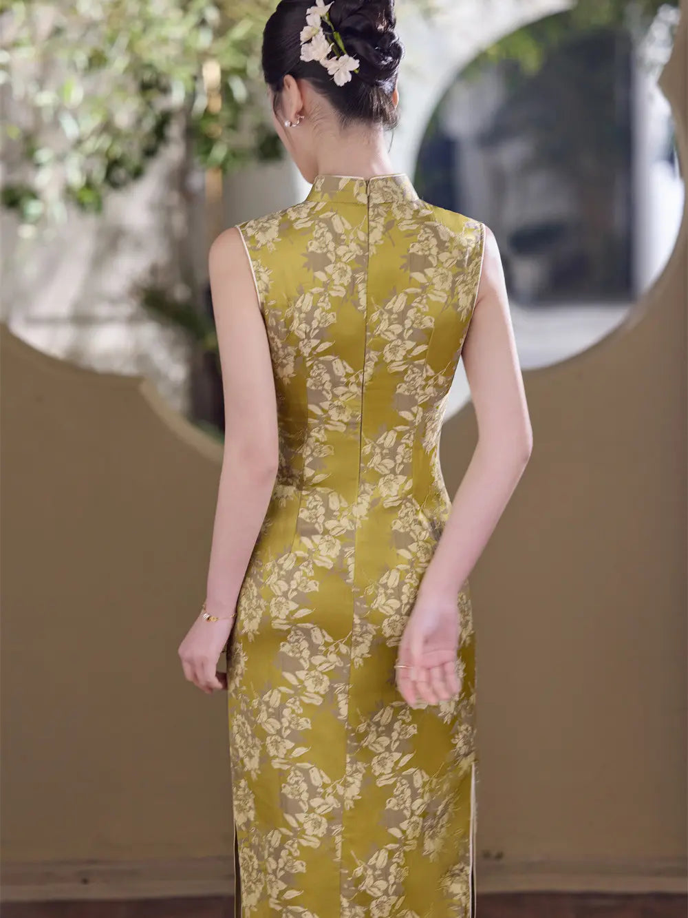 A woman viewed from behind wearing a sleeveless, floor-length cheongsam dress in olive green with a beige floral pattern. She has her hair styled in an updo with a small white flower accessory.