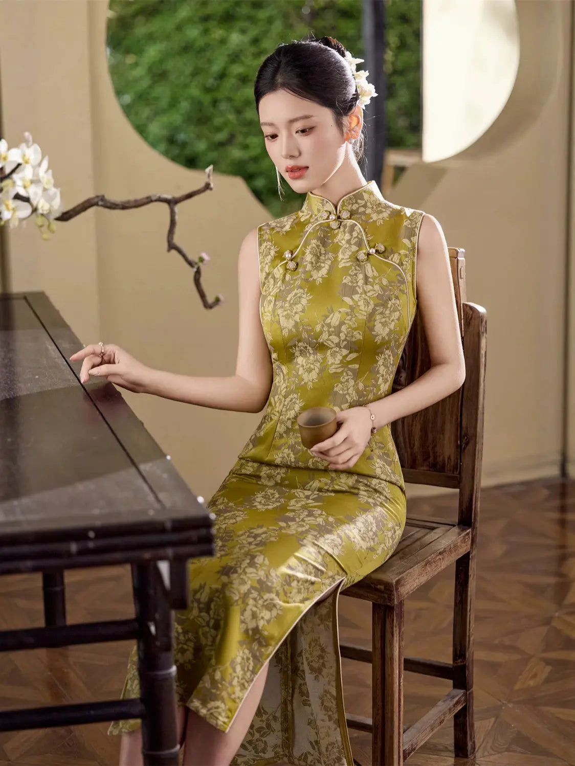 A woman seated on a wooden chair in a traditional Chinese-style interior, wearing a sleeveless olive-green cheongsam dress with a beige floral pattern and holding a small cup.