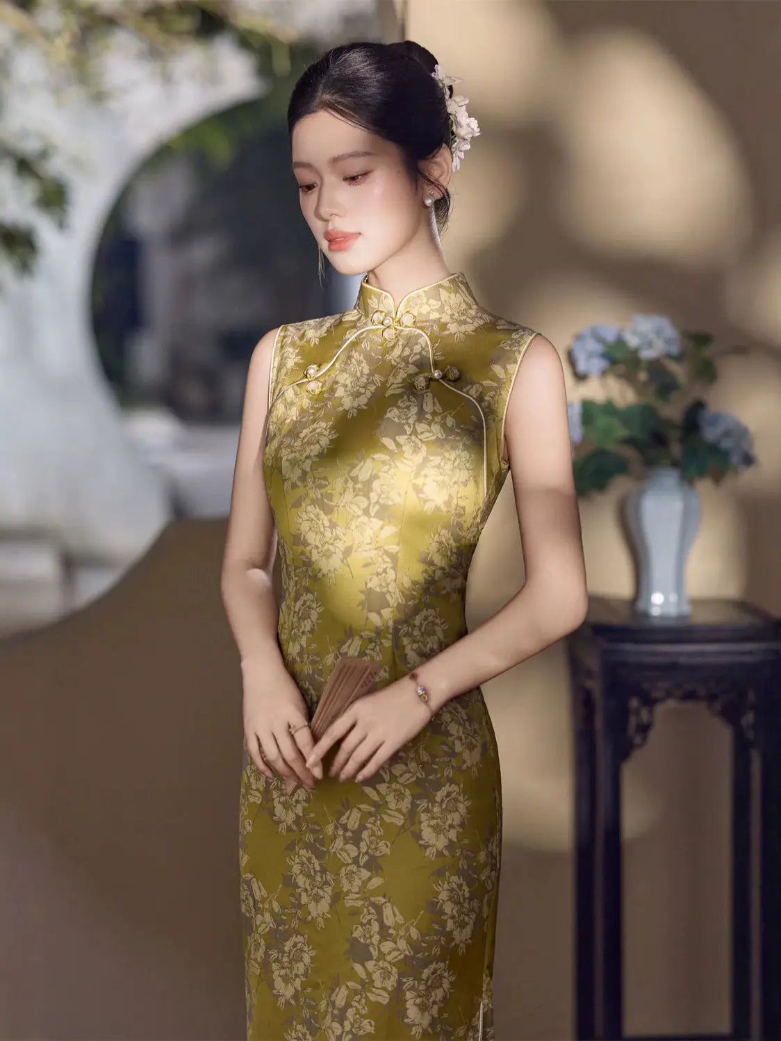 A woman wearing a sleeveless olive green and gold floral-patterned cheongsam (qipao) dress, standing in a softly lit indoor setting with a dark wooden table and flowers in the background.