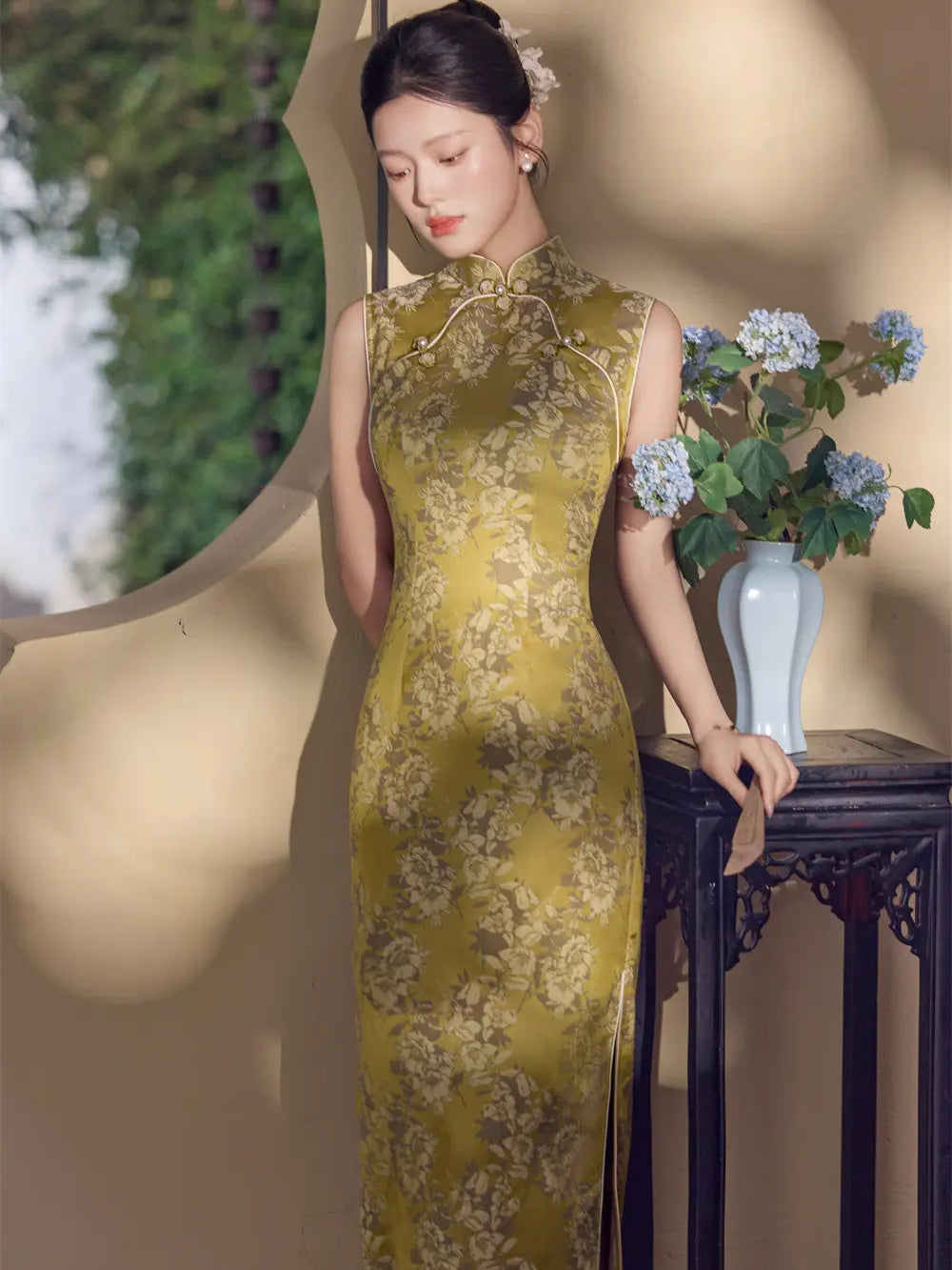 A woman wearing a sleeveless olive-yellow floral cheongsam dress stands beside a dark wooden table holding a vase of blue flowers, with warm sunlight and shadows across a beige wall.