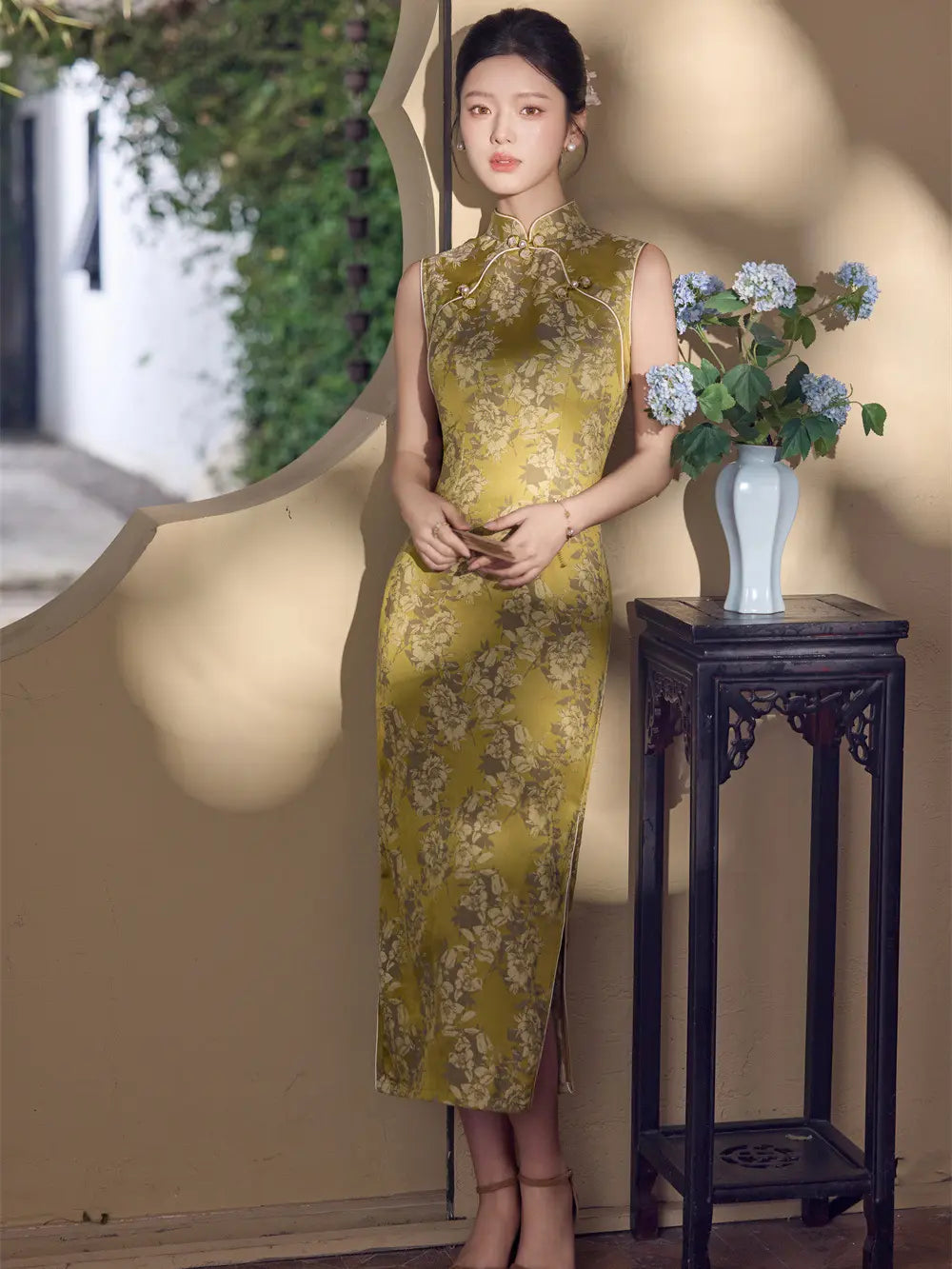 A woman stands indoors wearing a sleeveless, high-neck, floor-length cheongsam/qipao dress in olive green with a floral brocade pattern. She is posed beside a dark wooden side table holding a white vase with blue flowers against a beige wall with soft shadows.