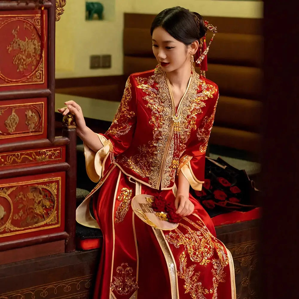 Woman in traditional red and gold Modernized V-Neck Chinese Wedding Xiu He Qun Kwa Dress  sitting next to a decorative wooden cabinet.