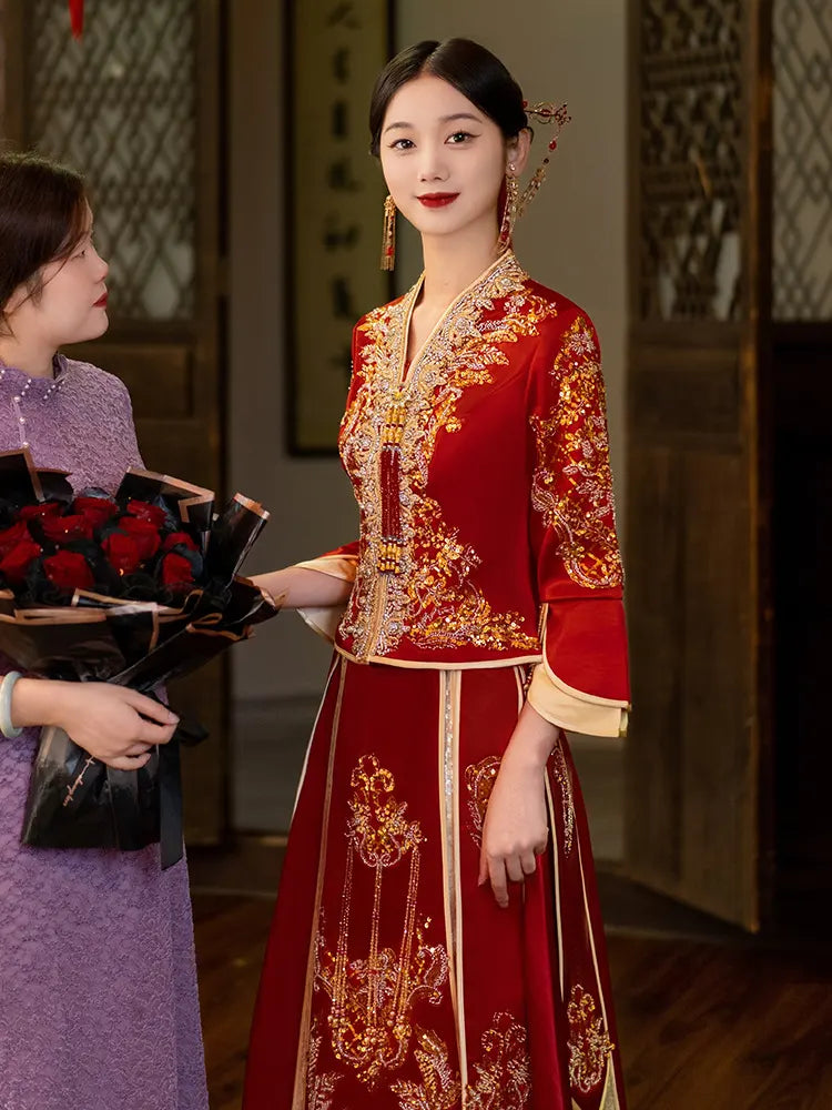 Woman in a red embroidered traditional Modernized V-Neck Chinese Wedding Xiu He Qun Kwa  outfit holding flowers, with another person in the background.