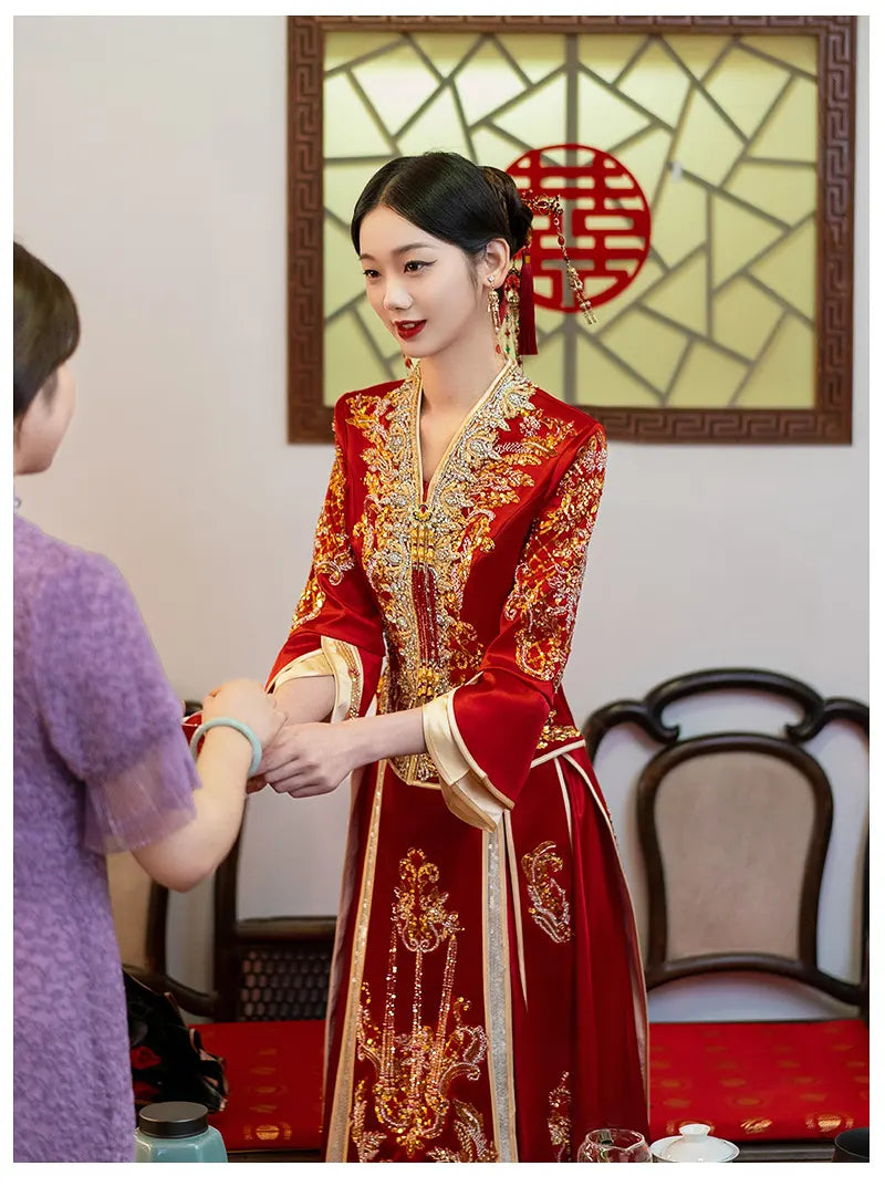 Woman in a traditional red and gold Modernized V-Neck Chinese Wedding Xiu He Qun Kwa Dress with intricate patterns, standing in a room with a decorative wall hanging.
