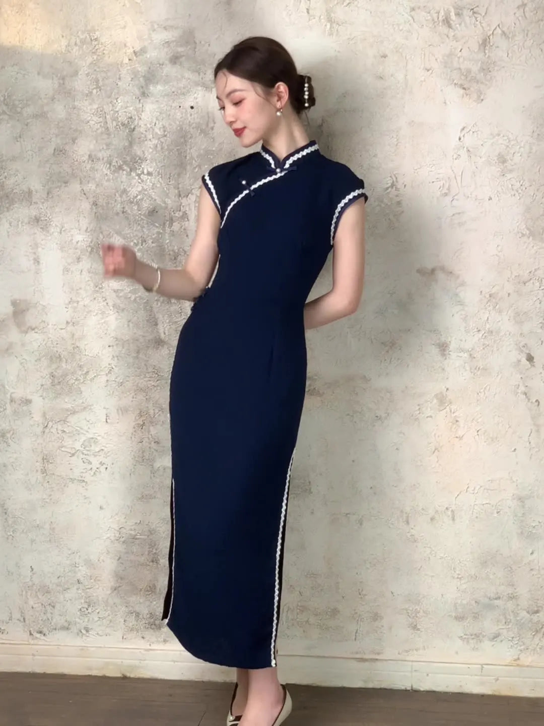Woman wearing a Navy Blue Cap Sleeves Jacquard Cotton Qipao Dress with white trim against a textured wall.