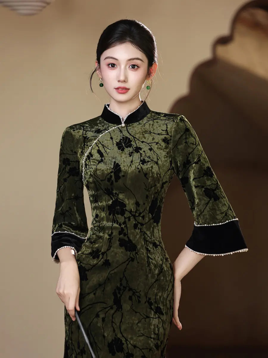 A woman modeling a dark olive green velvet cheongsam dress with black floral patterns, black collar and cuff trim, and silver bead accents, set against a warm beige background.