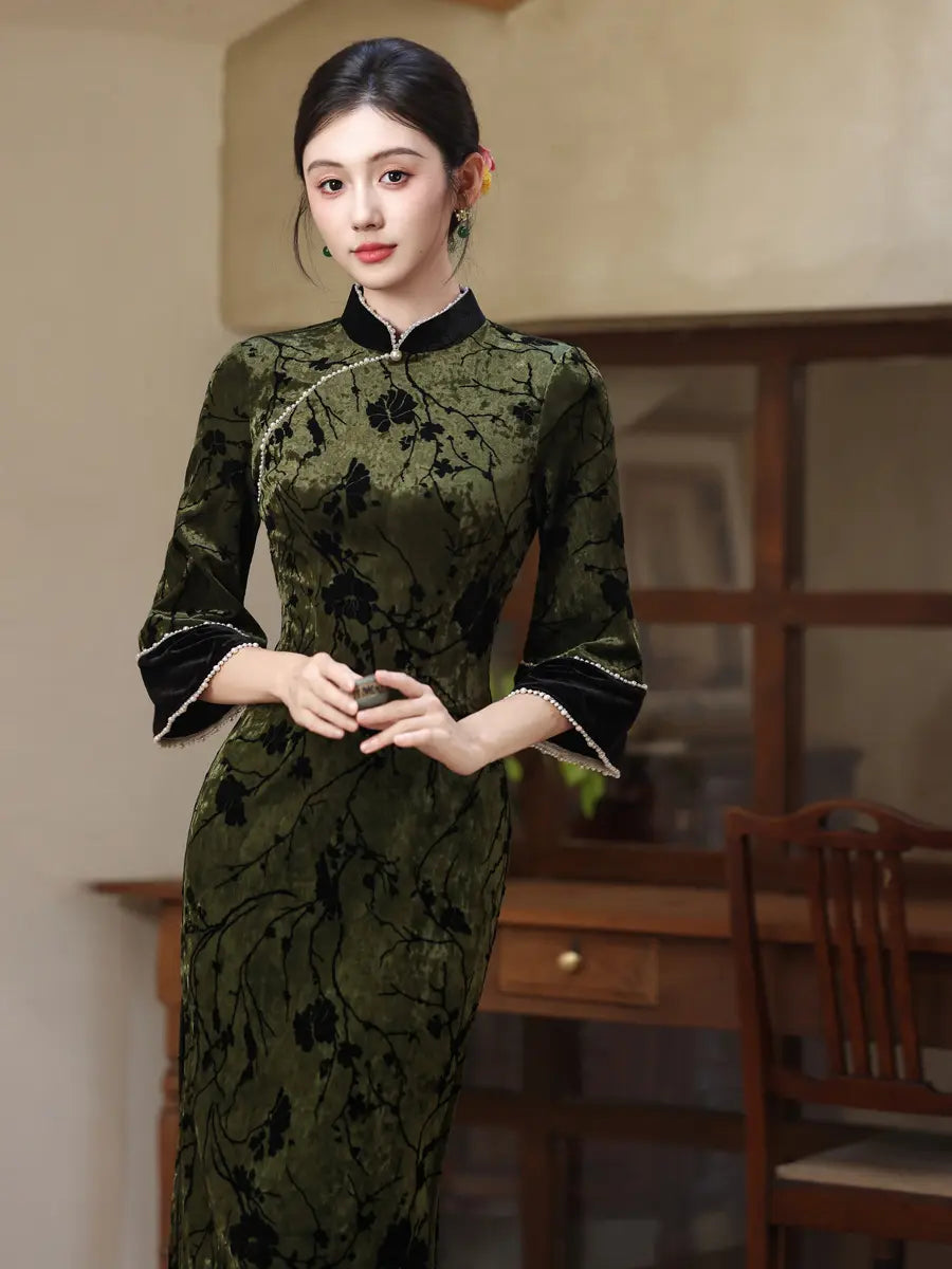 A woman wearing a dark olive green and black floral-patterned cheongsam dress with black trim and silver piping, standing in a warm-toned indoor room.