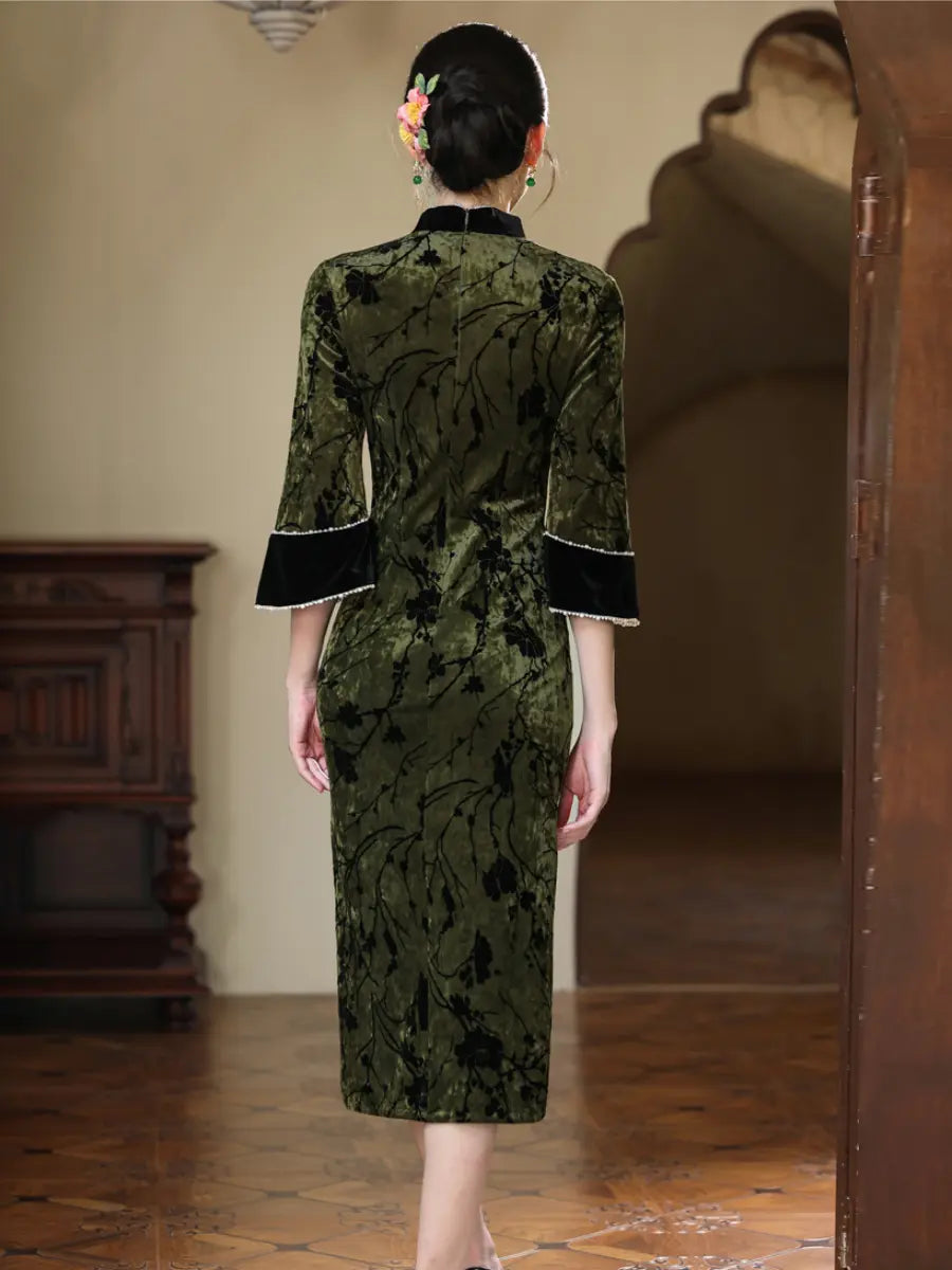 A woman viewed from behind wearing a dark green velvet cheongsam dress with a black floral pattern, black trim on the collar and cuffs, and a black skirt. She is standing in a traditional Chinese interior with wooden floors and arches.
