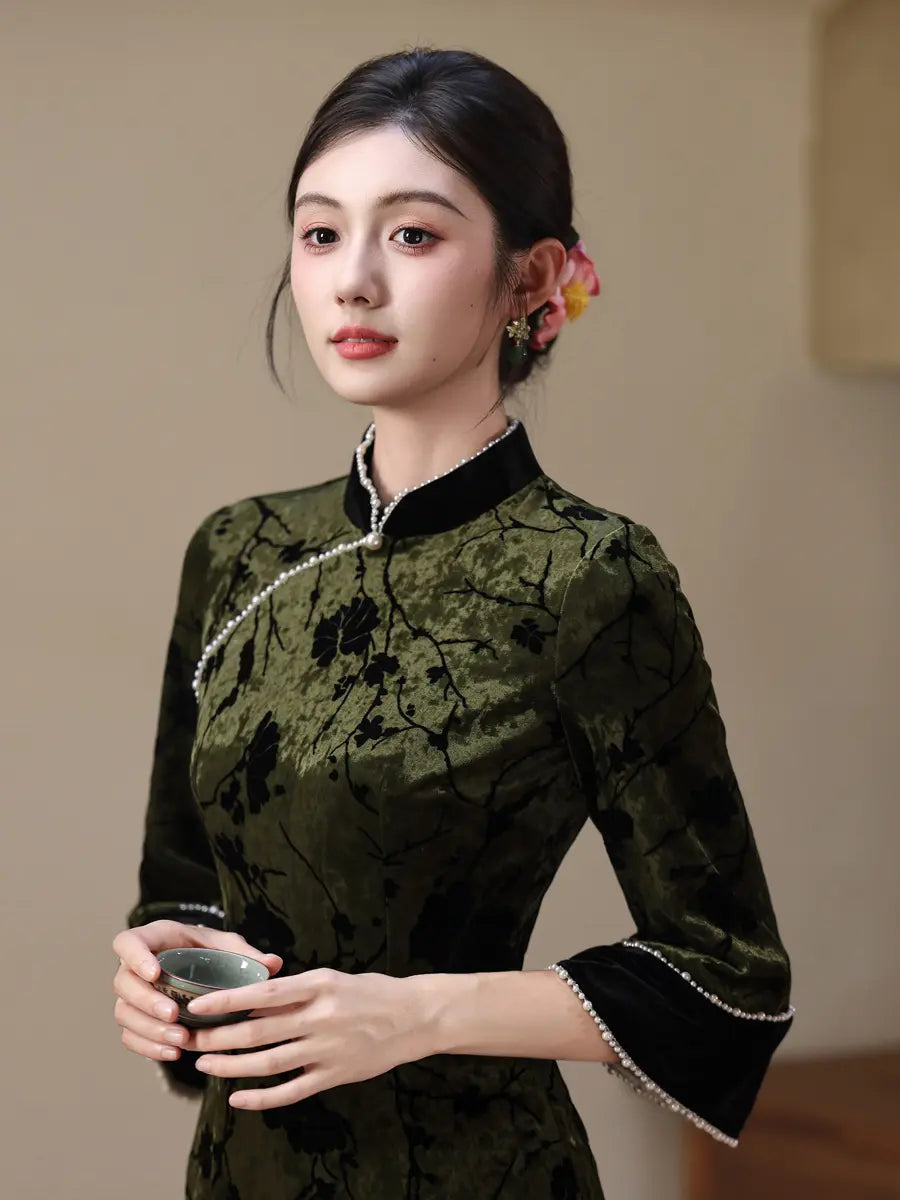 A woman wearing a dark green velvet cheongsam-style dress with black floral embroidery, black collar and cuffs, and white bead trim, holding a small cup. She has a pink and yellow flower in her hair against a neutral beige background.