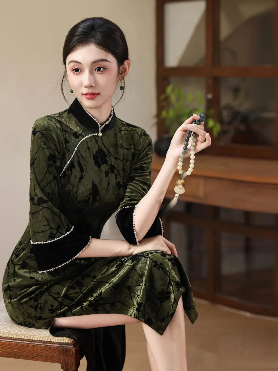 A woman seated indoors wearing a dark green and black patterned cheongsam-style dress with black trim and a beaded necklace, holding a beaded strand in one hand.