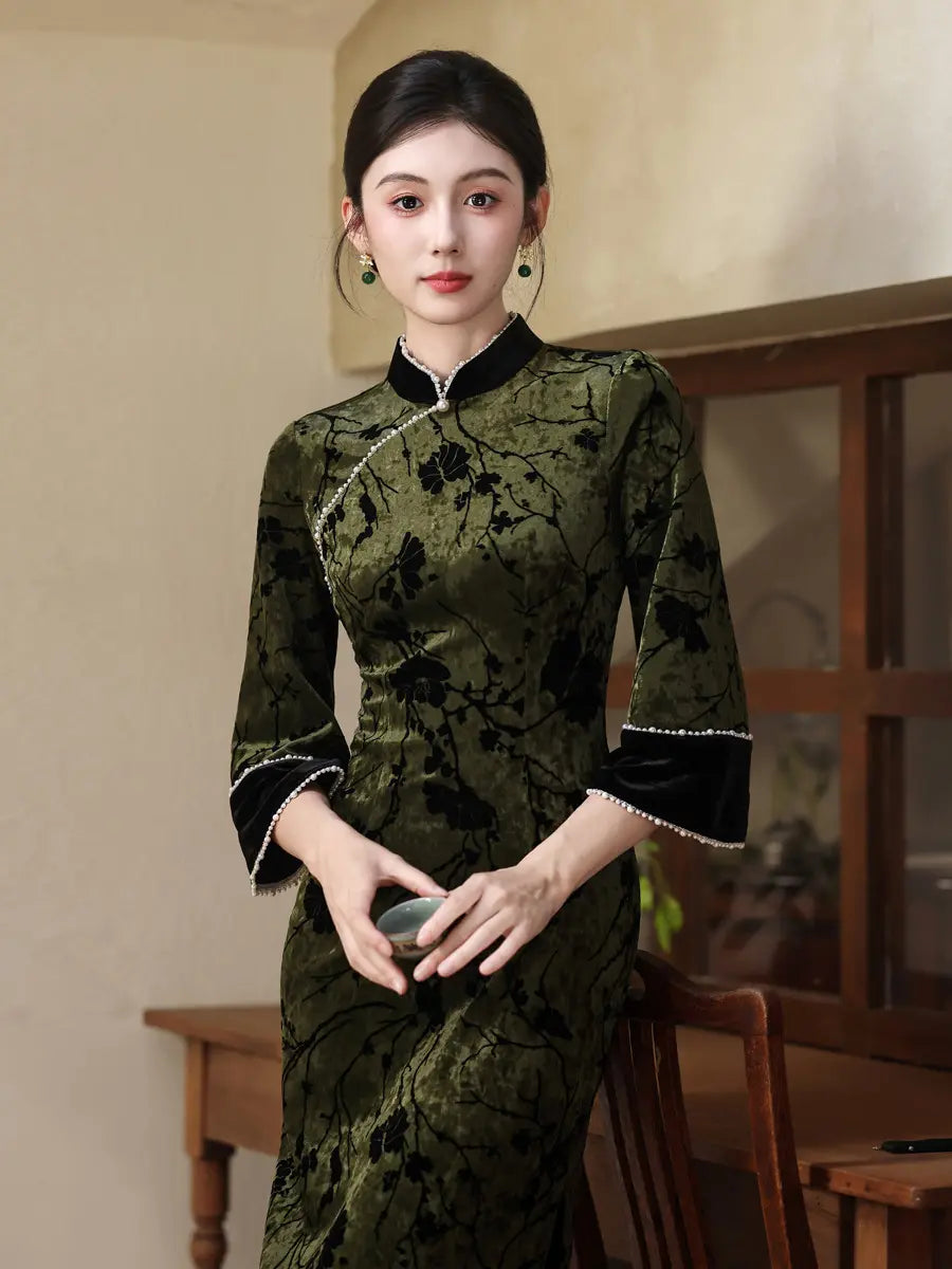 A woman wearing a dark olive green velvet cheongsam with black floral embroidery, black trim on the collar and cuffs, and white pearl accents, standing in a warm-toned indoor setting.