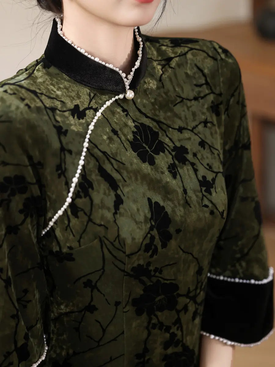 A close-up of a woman wearing a dark olive green and black floral embroidered cheongsam-style top with black velvet collar and cuff trim, accented by a pearl necklace and pearl trim.
