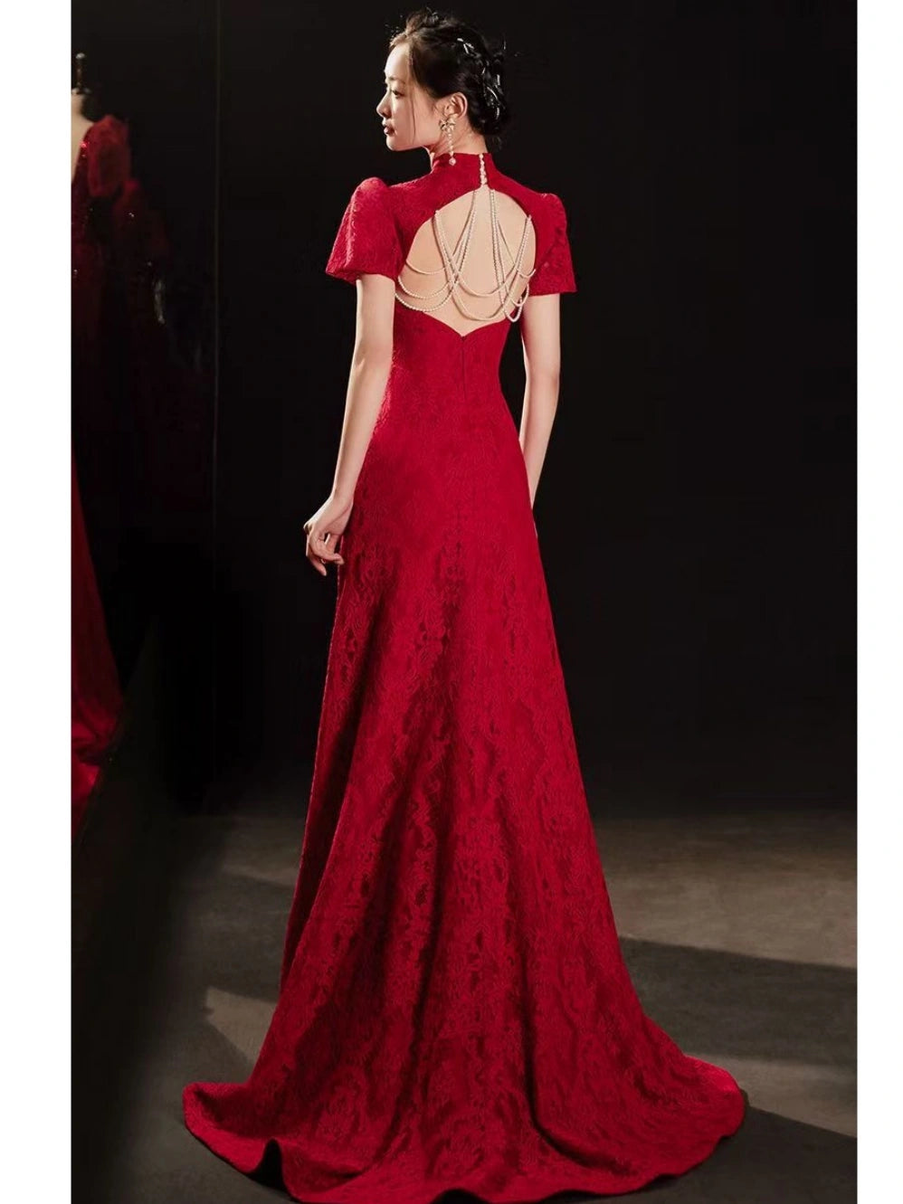 A woman modeling a floor-length red Open Back Lace Cheongsam/Qipao Dress With Train  evening gown from the back, featuring short puff sleeves, a high neckline, and an open back with layered chain detailing, set against a dark studio background.