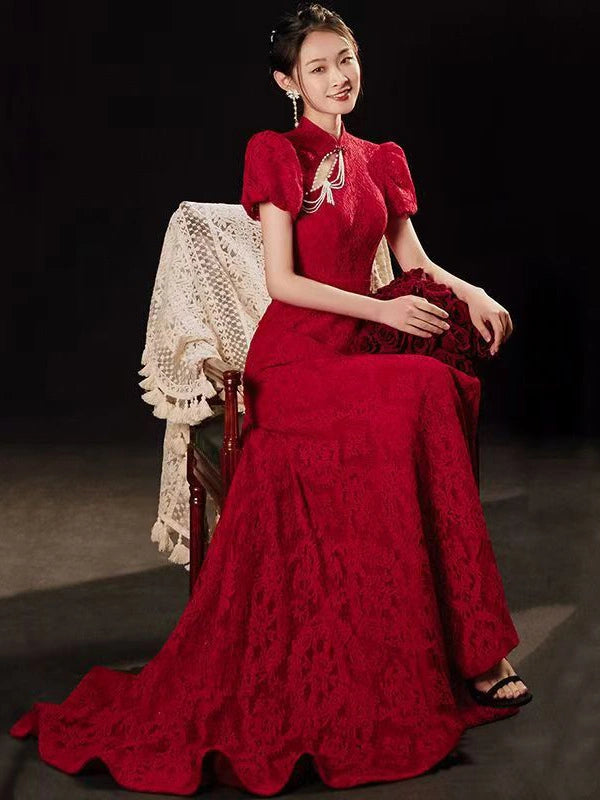 A woman seated on a dark wooden chair wearing a long red Open Back Lace Cheongsam/Qipao Dress With Train with short puff sleeves, a high neckline, and a train, against a dark studio background.