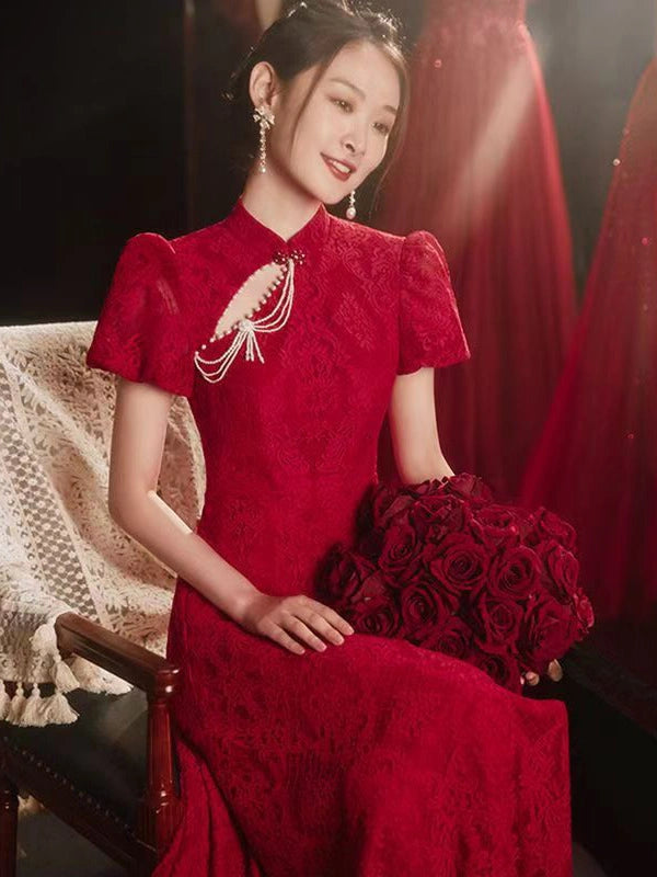 A woman seated on a dark wooden chair wearing a red lace cheongsam-style dress with short puff sleeves, a high collar, and a decorative pearl-like chest detail. She holds a bouquet of red roses in a dimly lit setting with red fabric in the background.