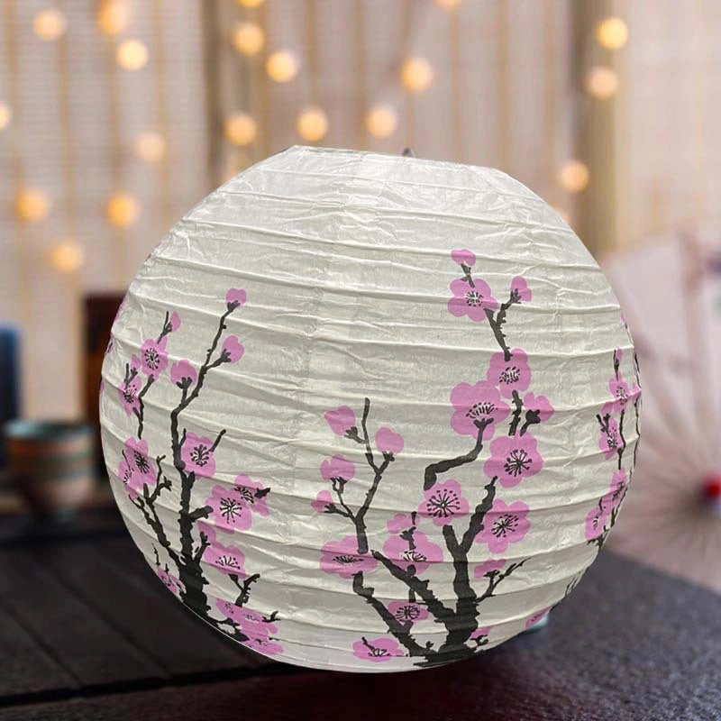 A round white paper lantern decorated with pink cherry blossom flowers and black branches, resting on a dark surface with a warm, blurred bokeh background.