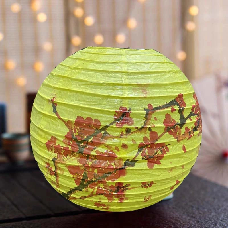 A round yellow paper lantern decorated with red cherry blossoms and dark branches, shown lit on a dark table with warm bokeh lights in the background.