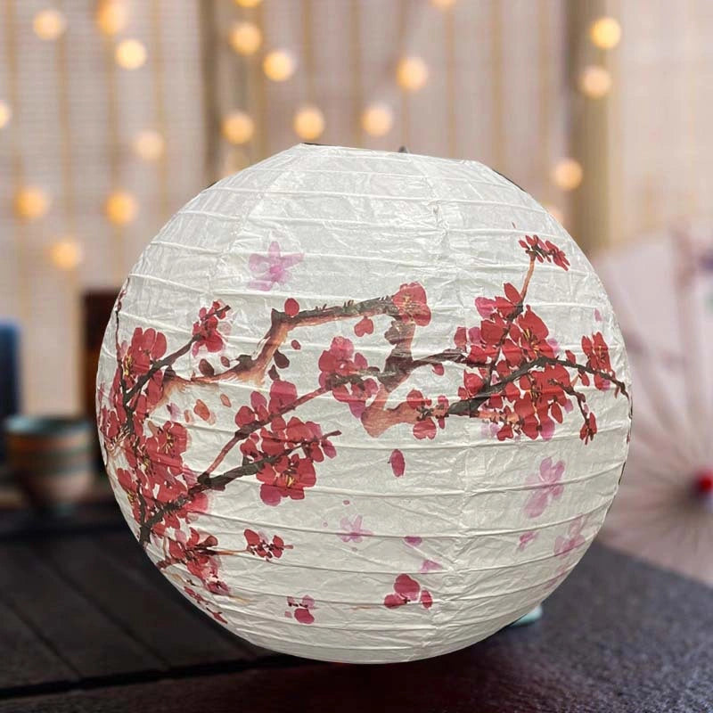A round white paper lantern decorated with a red and pink cherry blossom branch design, resting on a dark table with warm bokeh string lights in the background.