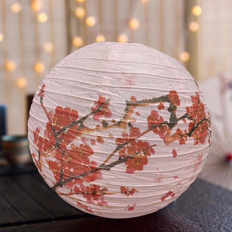 A round pink Chinese paper lantern decorated with a red cherry blossom branch pattern, resting on a dark surface with a warm, blurred bokeh background.