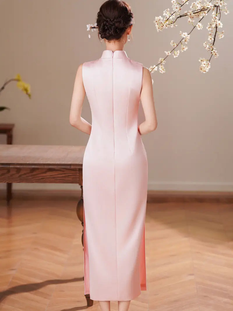 A woman stands with her back to the camera wearing a sleeveless light pink cheongsam-style dress with a high mandarin collar and side slit, photographed indoors against a neutral wall with wooden flooring and flowering branches in the upper right.