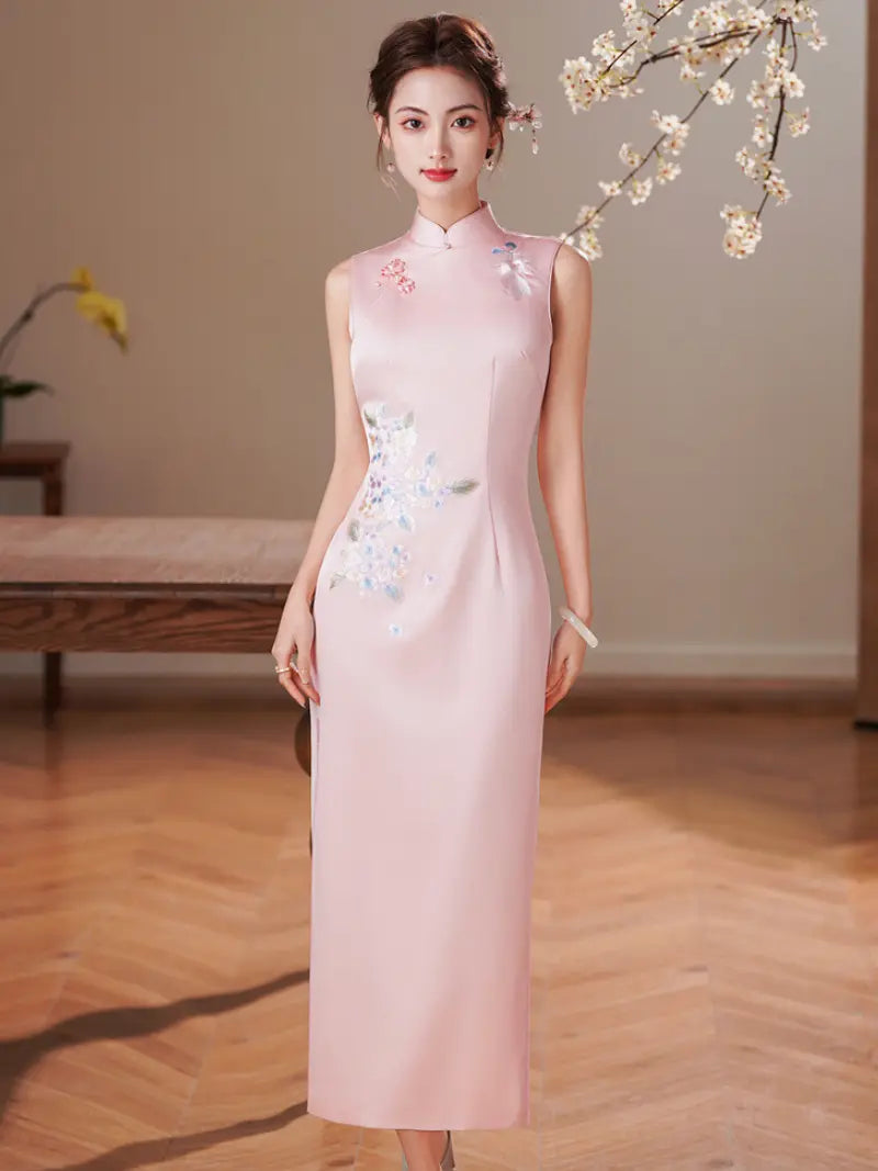 A woman wearing a sleeveless light pink cheongsam-style dress with floral embroidery on the chest and hip, standing in a warm indoor setting with wooden flooring and decorative branches overhead.