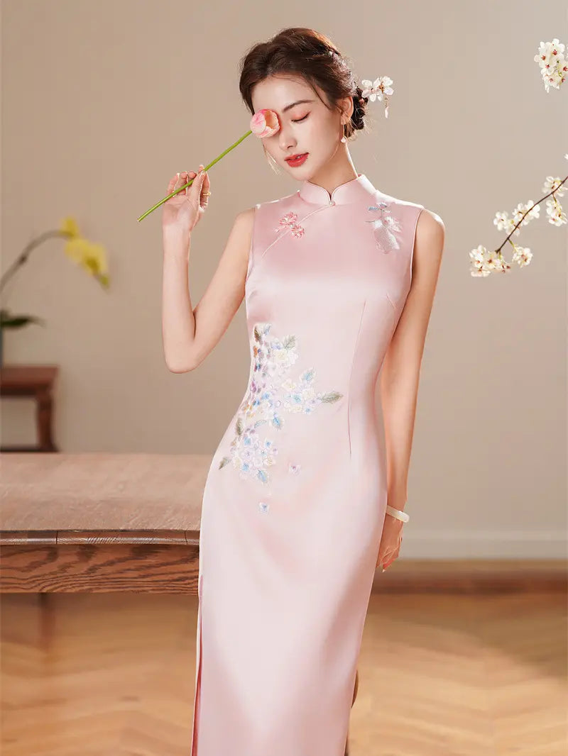 A woman wearing a sleeveless light pink cheongsam with floral embroidery, holding a pink flower in front of her eye, in a warm-toned indoor setting.