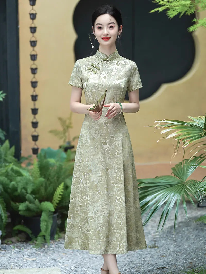 A woman stands outdoors wearing a short-sleeve, midi-length Pistachio Sage Green Floral Jacquard Modern A Line Qipao cheongsam-style dress in a light beige or champagne tone with an allover tonal floral jacquard pattern. The dress has a high collar, a self-tie neckline, and a decorative floral brooch on the chest.