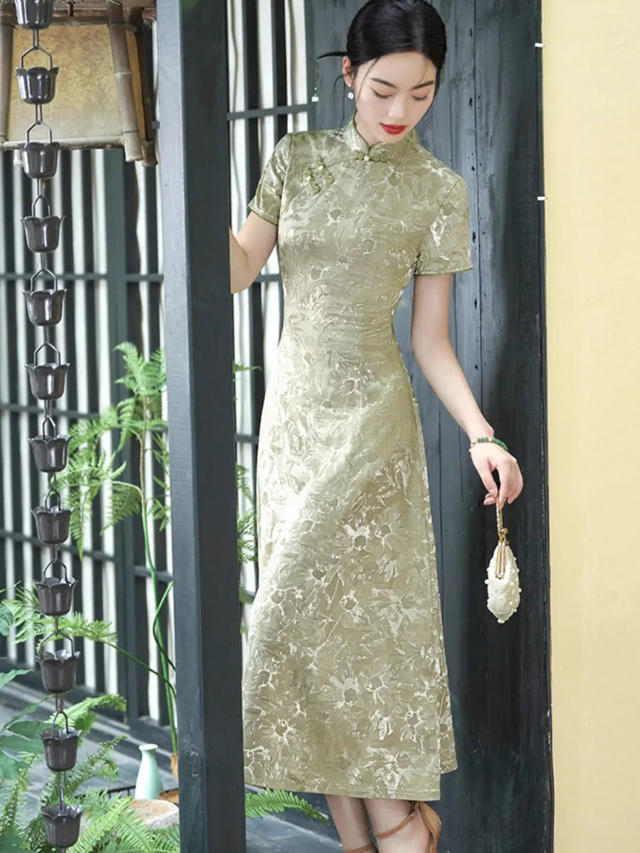 A woman wearing a short-sleeved, knee-length Pistachio Sage Green Floral Jacquard Modern A Line Qipao cheongsam dress in a shimmering champagne-gold floral jacquard pattern, standing in front of a dark wooden door with a hanging lantern and plants.