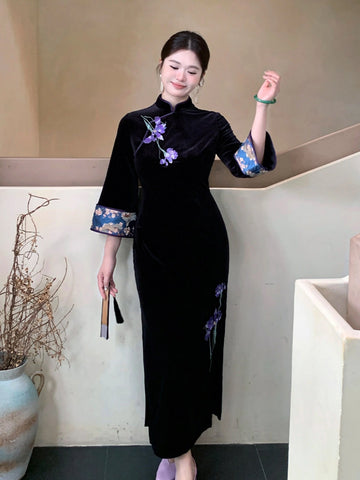 A woman wearing a black velvet cheongsam dress with purple floral embroidery and blue patterned cuffs, standing in an indoor setting with a beige wall and staircase.