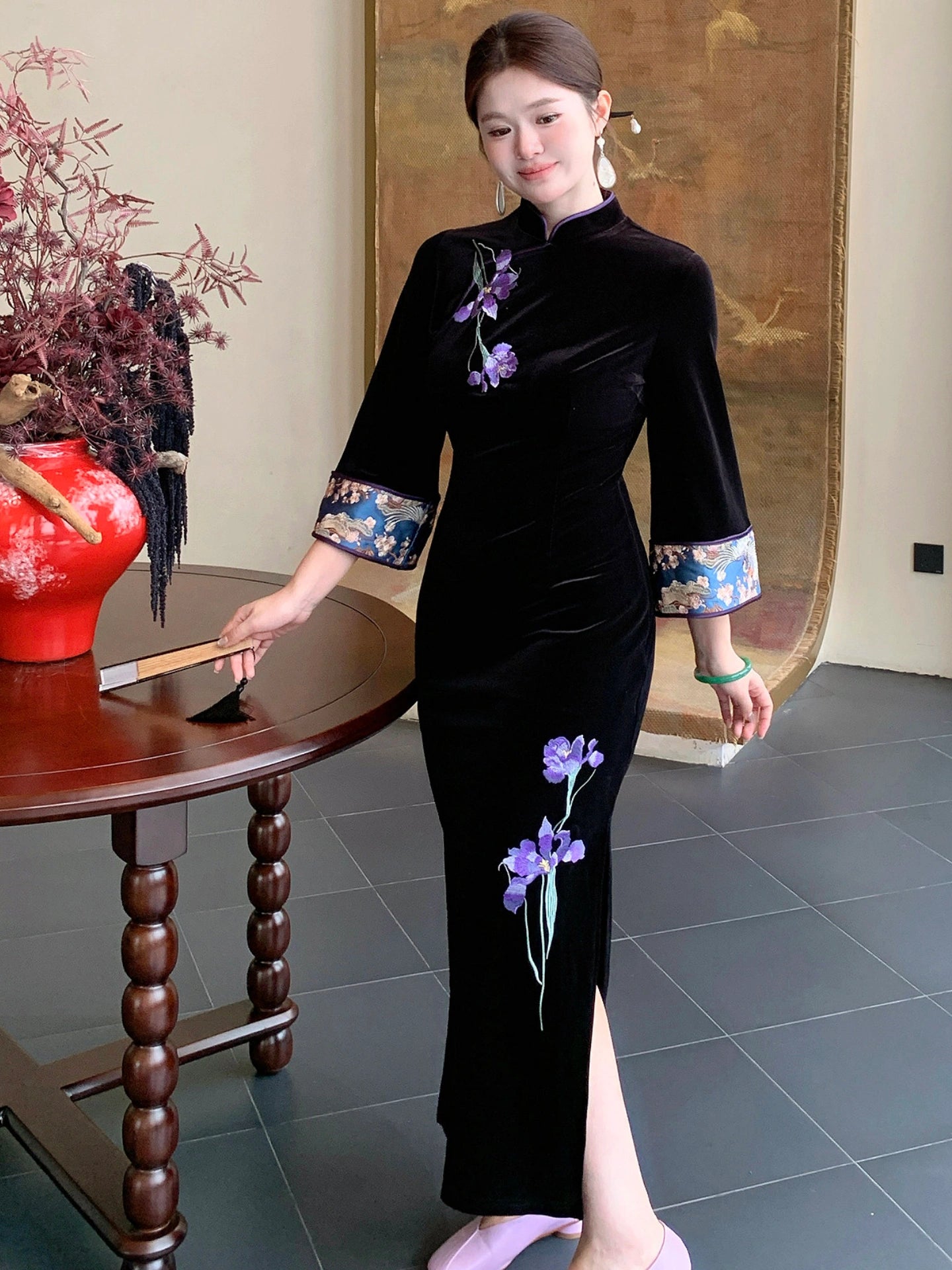 A woman wearing a black velvet cheongsam dress with purple floral embroidery and patterned blue and gold cuffs, standing in a traditional indoor setting next to a wooden table.