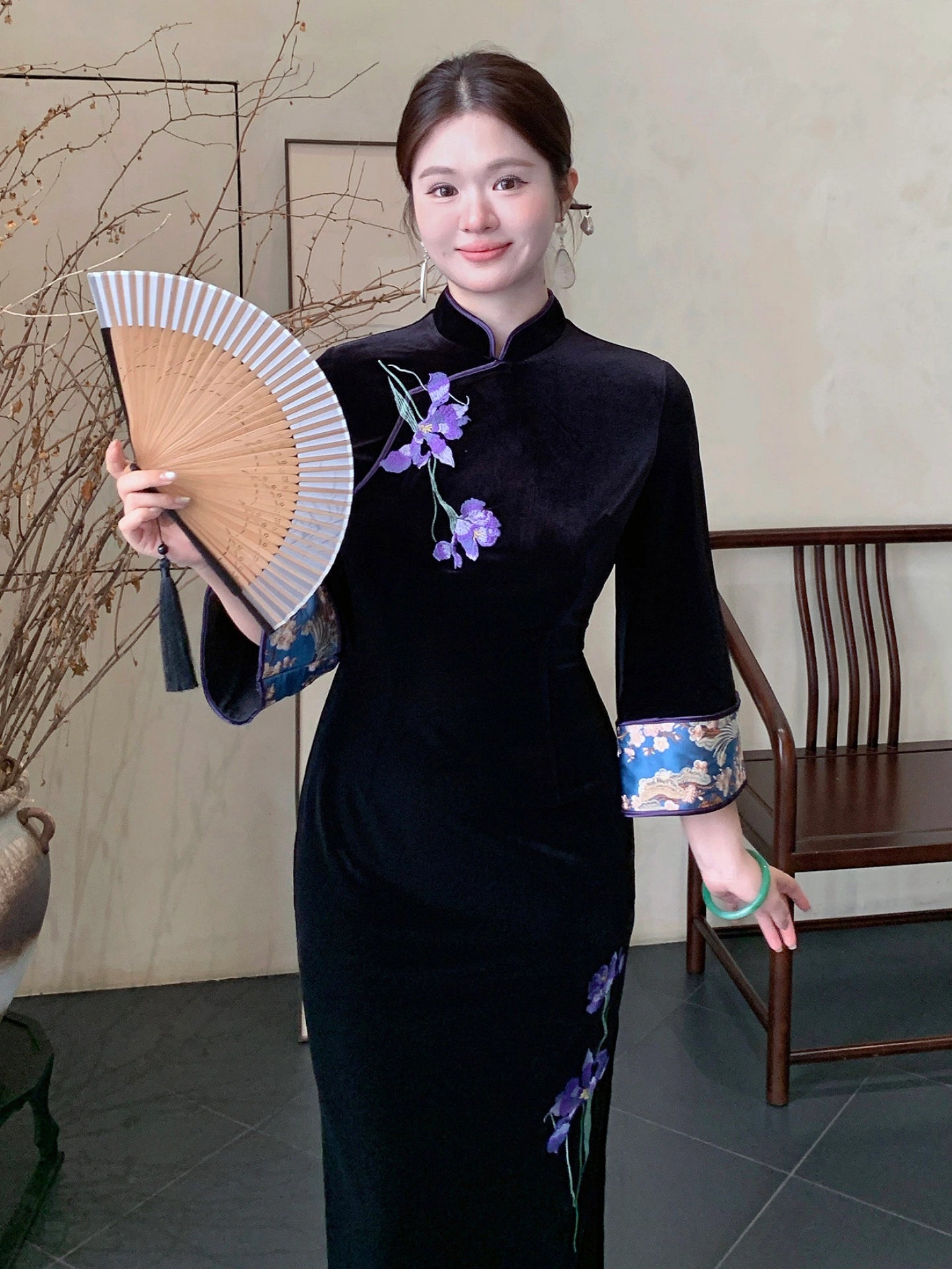 A woman wearing a black velvet cheongsam-style dress with purple floral embroidery and blue patterned cuffs, holding a folding fan in a traditional indoor setting.