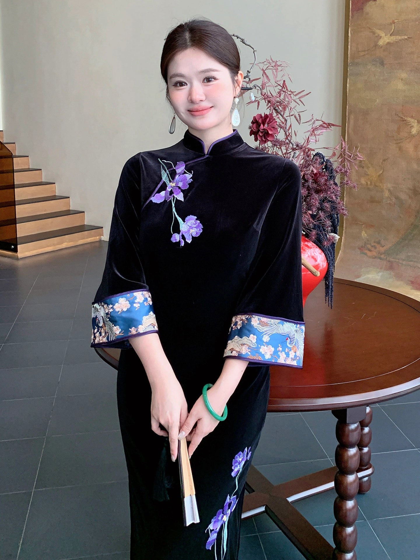 A woman wearing a black velvet cheongsam with purple floral embroidery and blue patterned cuffs, holding a folding fan in a traditional indoor setting.