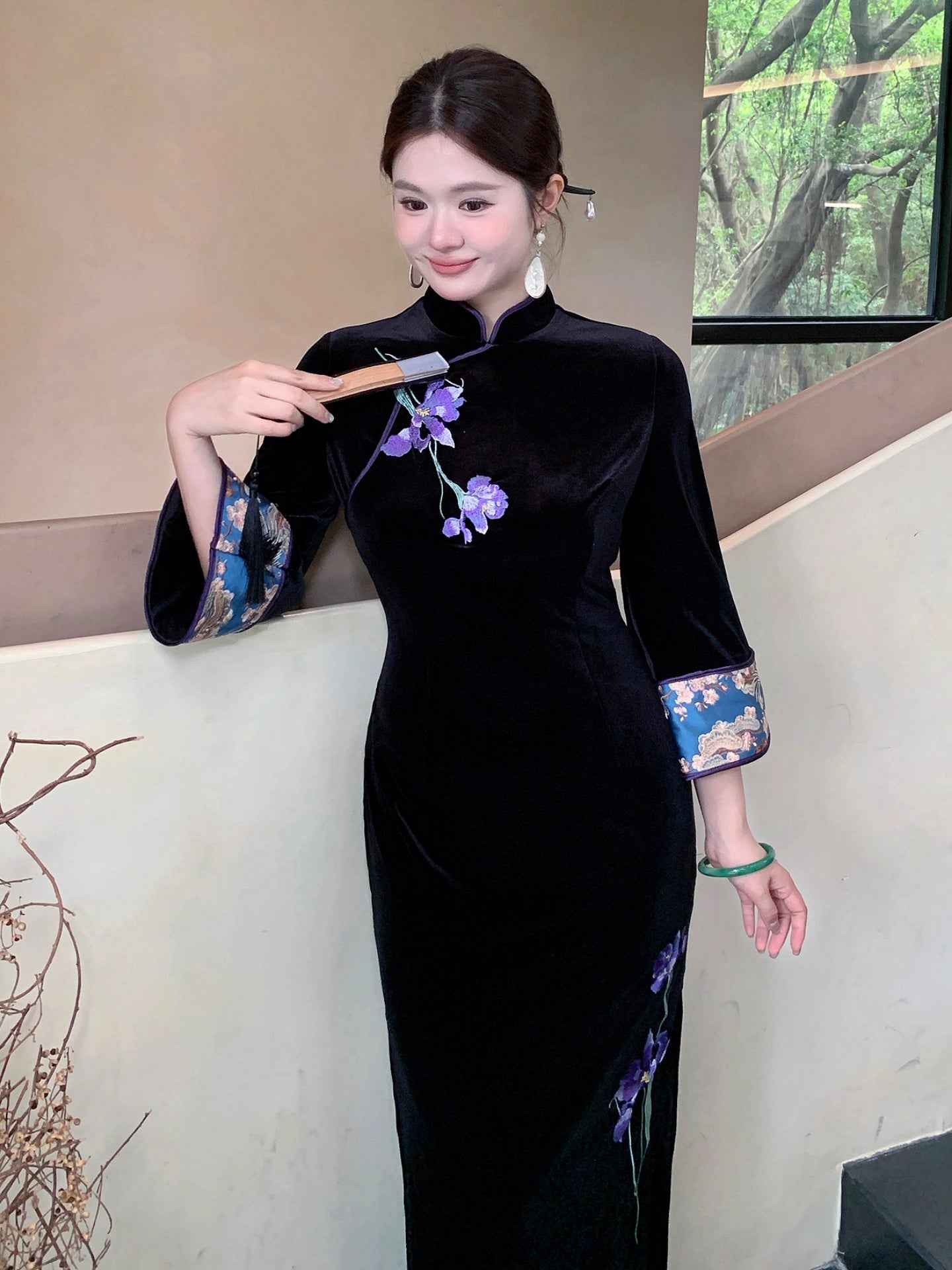 A woman wearing a black velvet cheongsam-style dress with purple floral embroidery and blue patterned cuff details, holding a folding fan in an indoor setting.