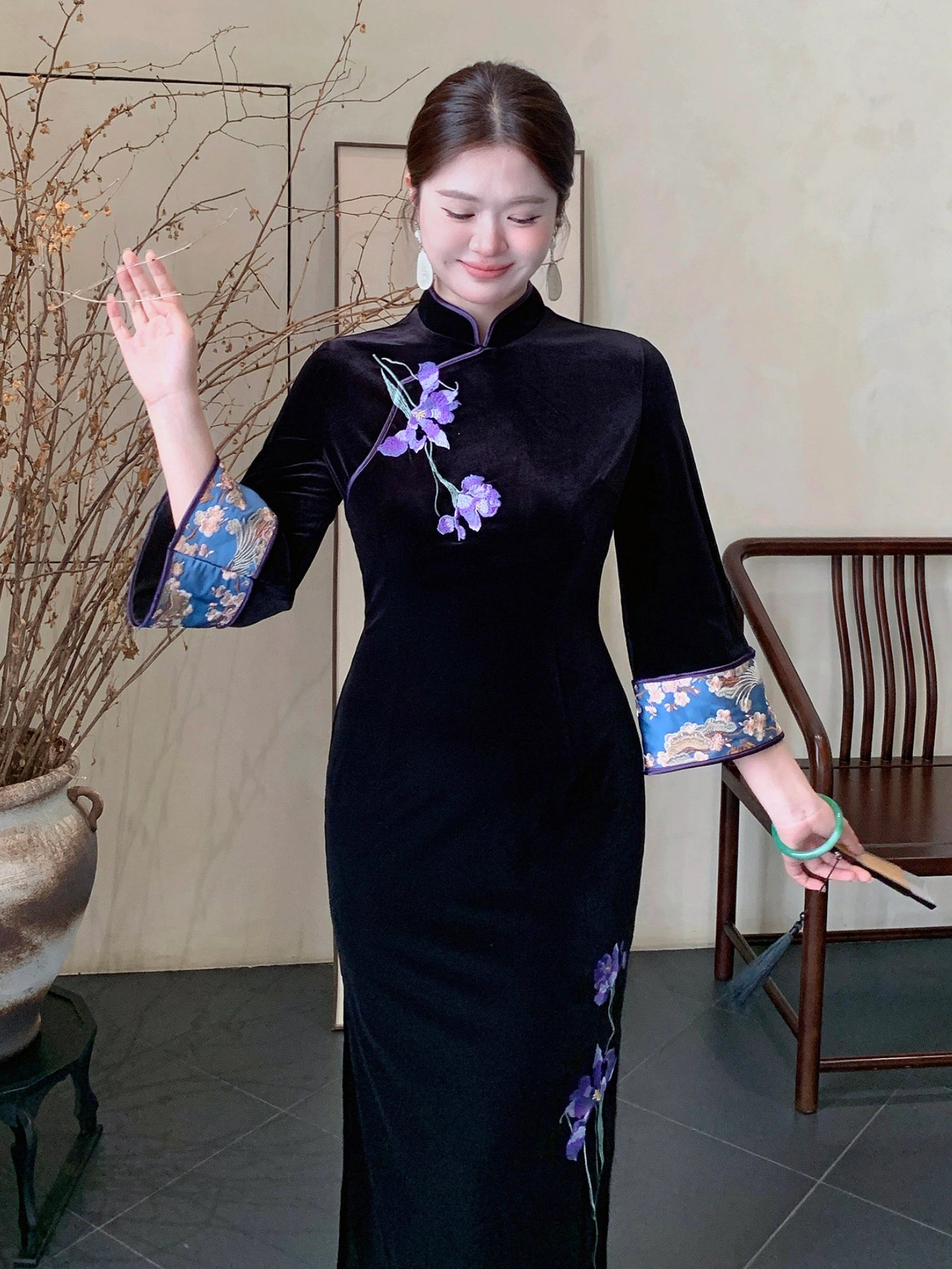 A woman wearing a black velvet cheongsam with purple floral embroidery and blue patterned cuffs, standing in a traditional indoor setting with a wooden chair and dried branches.