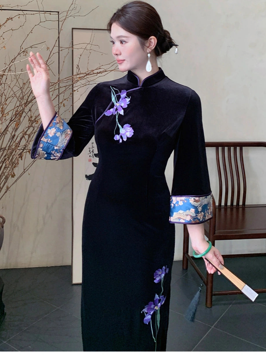 A woman wearing a black velvet cheongsam dress with purple floral embroidery and blue patterned sleeve cuffs, standing in a traditional indoor setting.