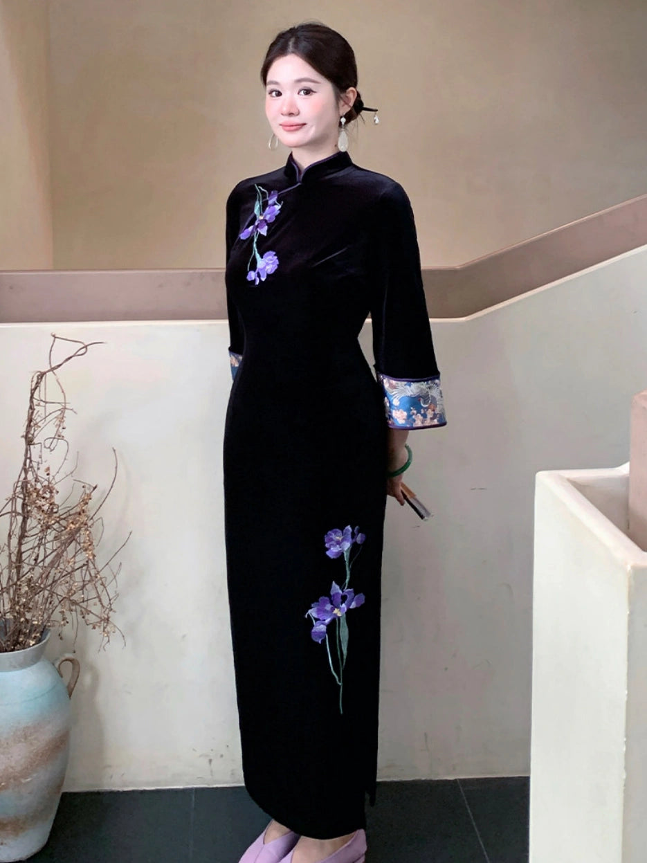 A woman wearing a black velvet cheongsam dress with purple floral embroidery and patterned cuffs, standing in an indoor setting.