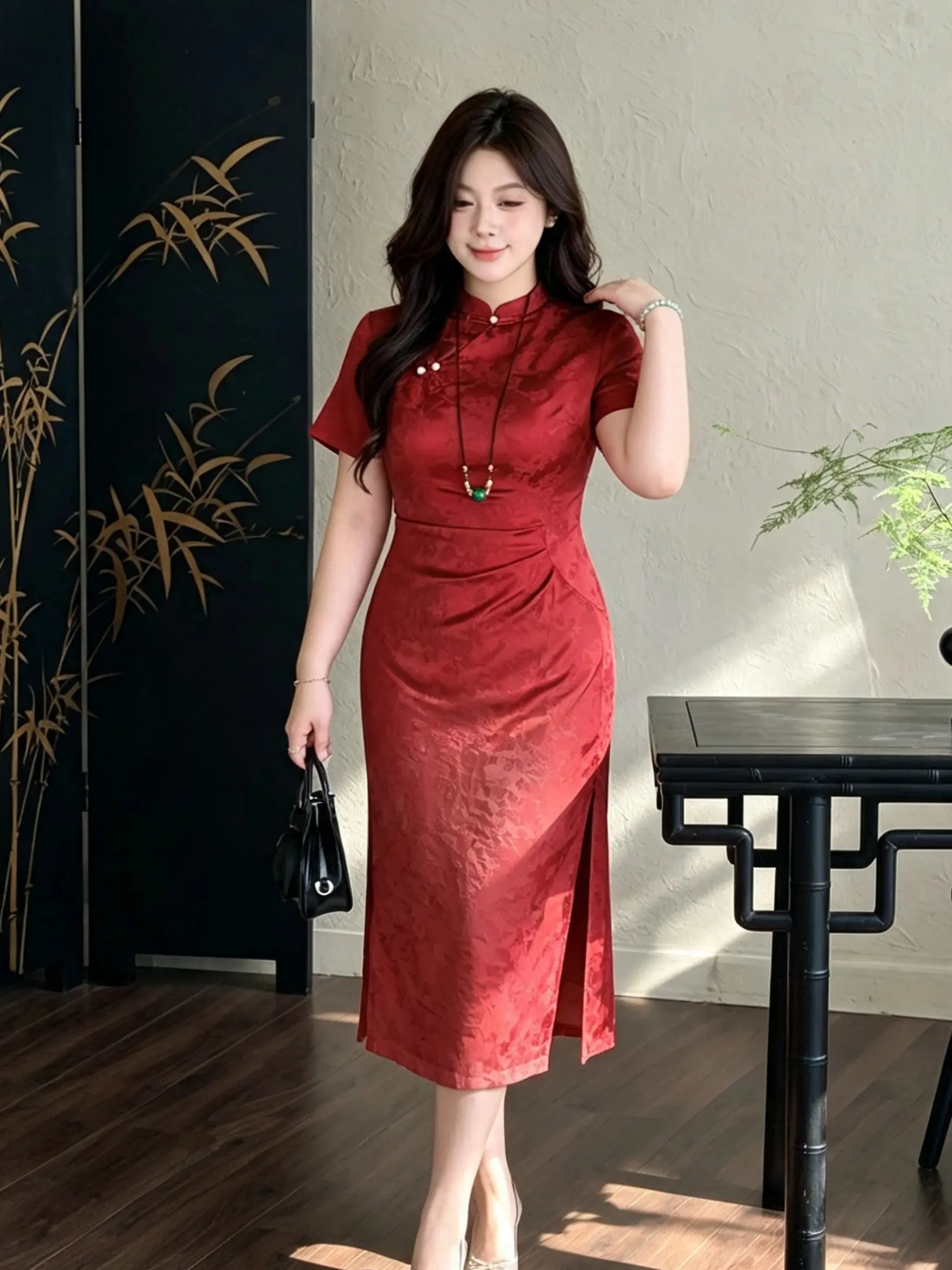A woman modeling a red short-sleeve midi cheongsam dress with a subtle tonal pattern, standing in a styled indoor setting with a black screen, table, and textured wall.