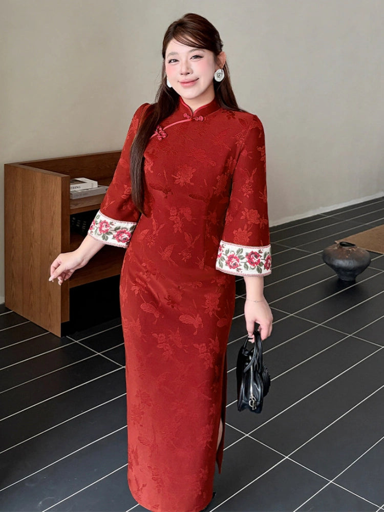 A woman wearing a Plus size red floral jacquard cheongsam/qipao dress with white floral-trimmed cuffs, standing in an indoor setting with a wooden cabinet and dark tiled floor.