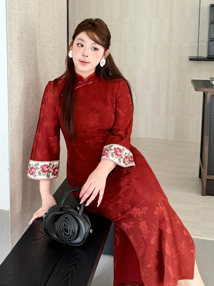 A woman wearing a red floral jacquard cheongsam dress with embroidered floral cuffs, posing on a dark wooden bench and holding a black rose-shaped handbag.