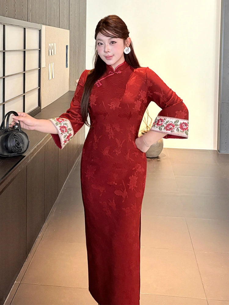 A woman wearing a red long-sleeve cheongsam dress with floral embroidery on the cuffs, standing indoors next to a counter.