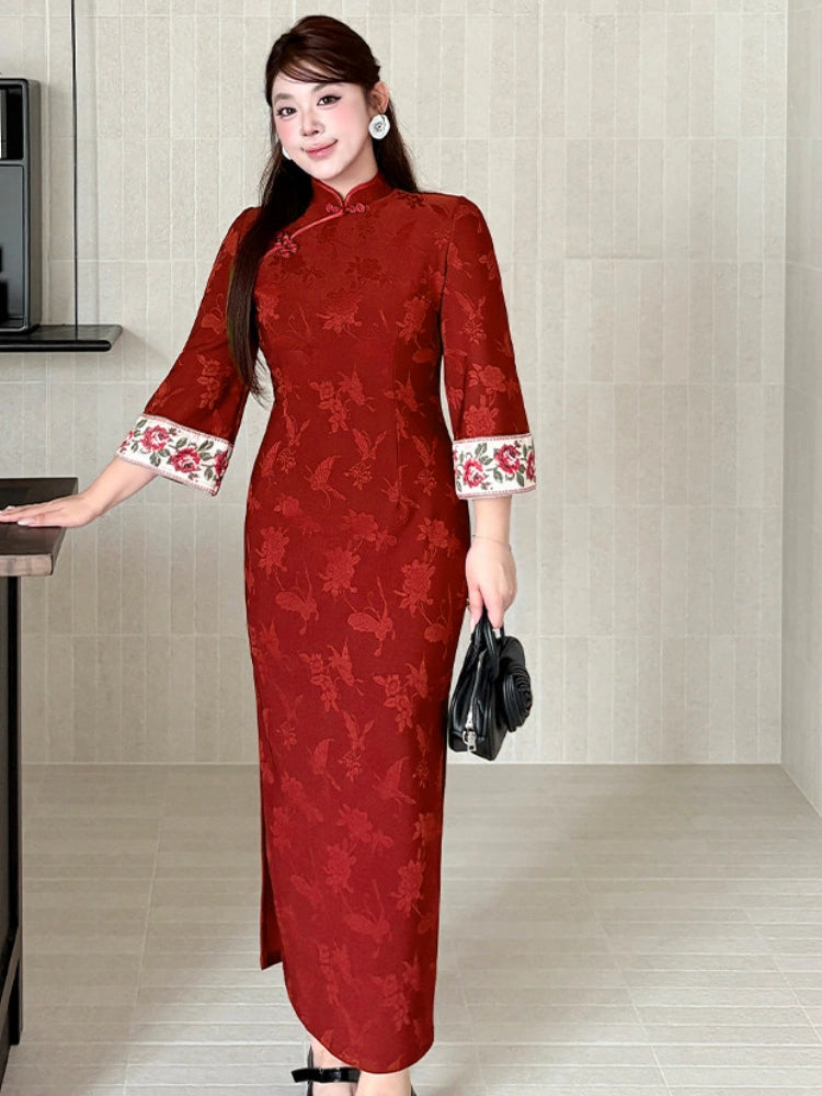 A woman modeling a red floral jacquard cheongsam dress with white cuffs featuring red floral embroidery, holding a black handbag in a modern tiled indoor setting.
