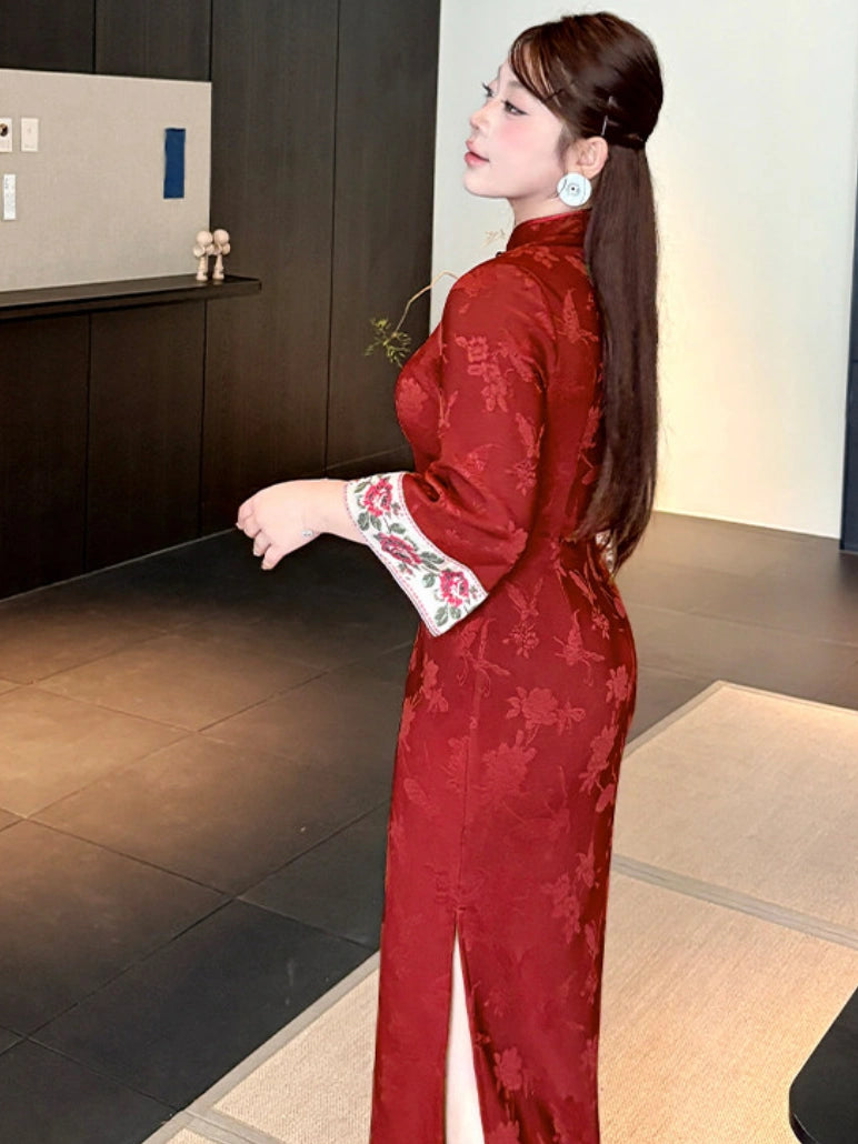 A woman wearing a red plus size Chinese cheongsam/qipao dress with floral embroidery on the sleeves, standing in a modern indoor setting.