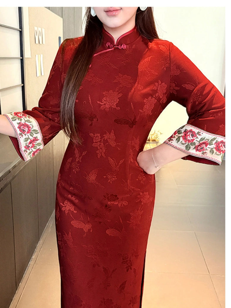 A woman wearing a red floral jacquard cheongsam/qipao dress with embroidered floral cuffs, standing indoors.