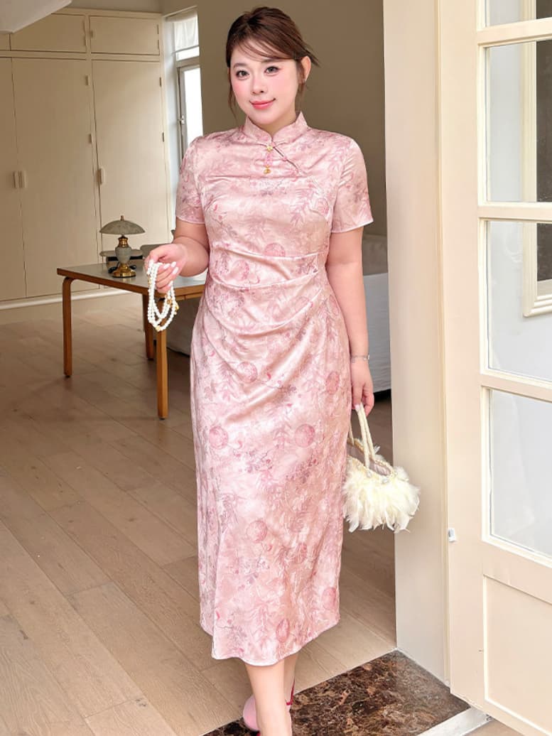 A woman wearing a pink floral-patterned short-sleeve plus size qipao cheongsam dress, holding a white fluffy handbag and a pearl necklace, standing in a doorway of a home interior.