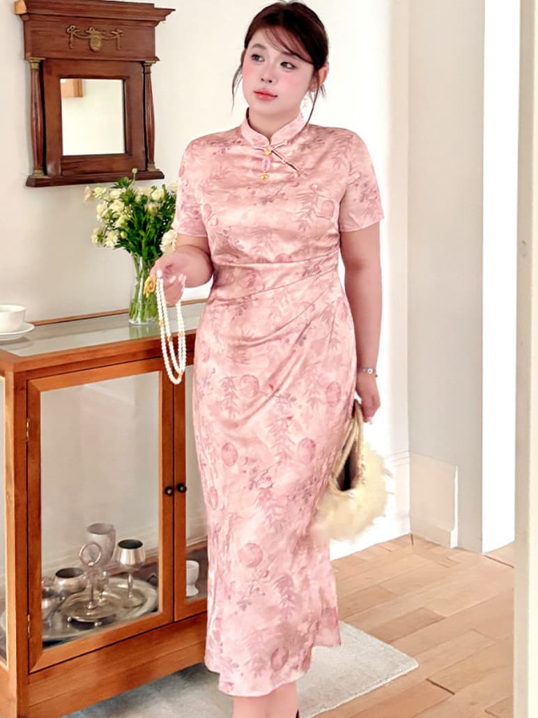 A woman wearing a pink short-sleeve plus size cheongsam dress with a tonal floral pattern, standing in a room with wooden furniture and holding a pearl necklace and a small handbag.