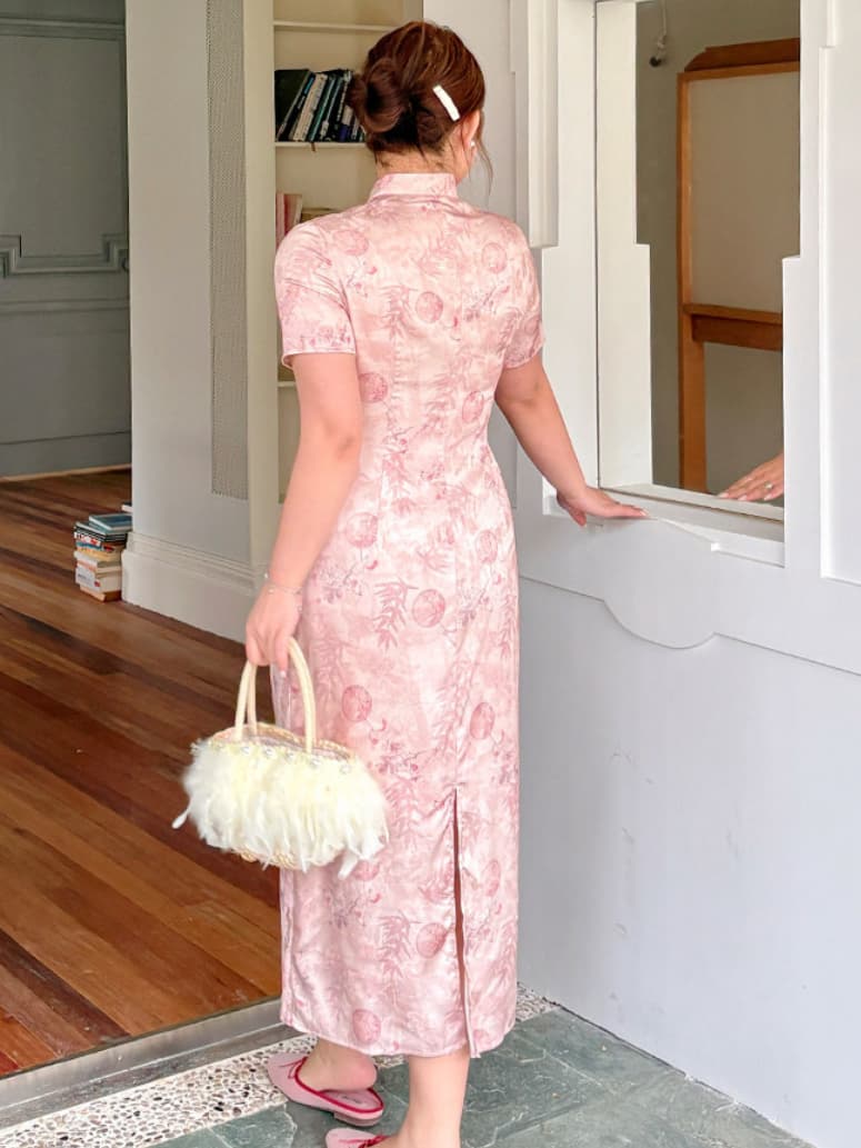 A woman stands with her back to the camera wearing a light pink floral-patterned plus size cheongsam-style dress, holding a fluffy cream handbag, in an indoor setting with a window and wooden floor visible.