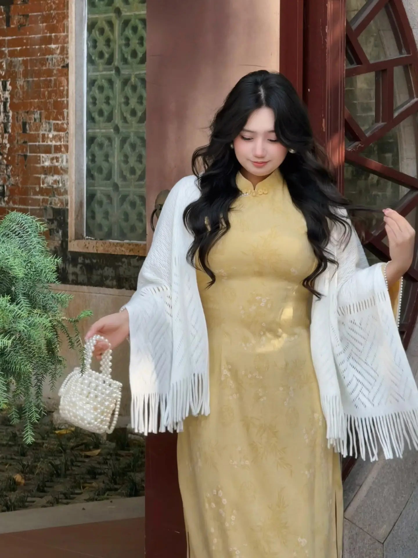 Woman in a Plus Size Golden Yellow Long Sleeve Jacquard Qipao with Lace Trim  dress with a white shawl holding a small handbag, standing in front of a textured wall.
