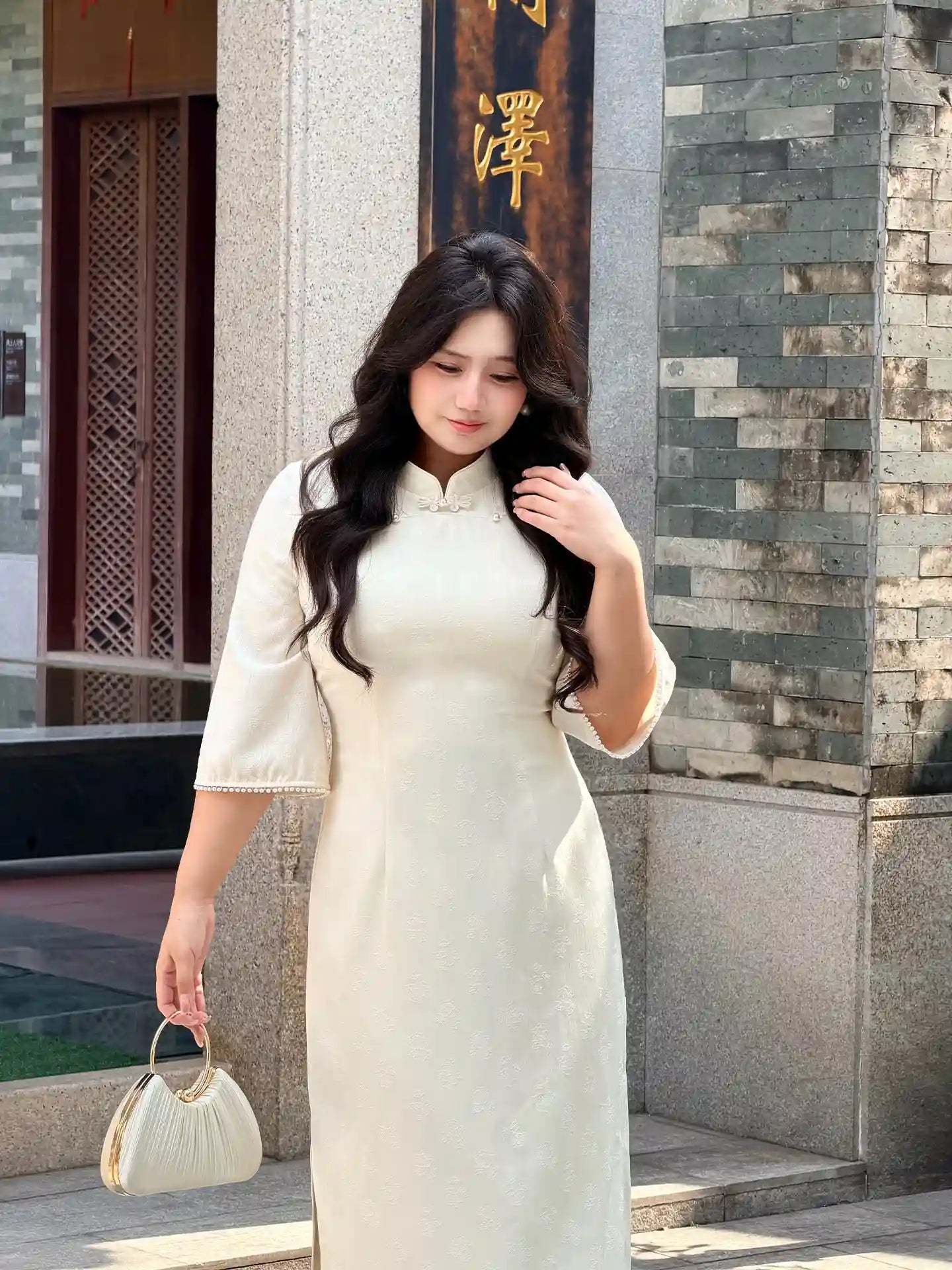 Woman in a Plus Size Ivory White Floral Long Sleeve Jacquard Qipao  standing in front of a stone wall with Chinese characters.