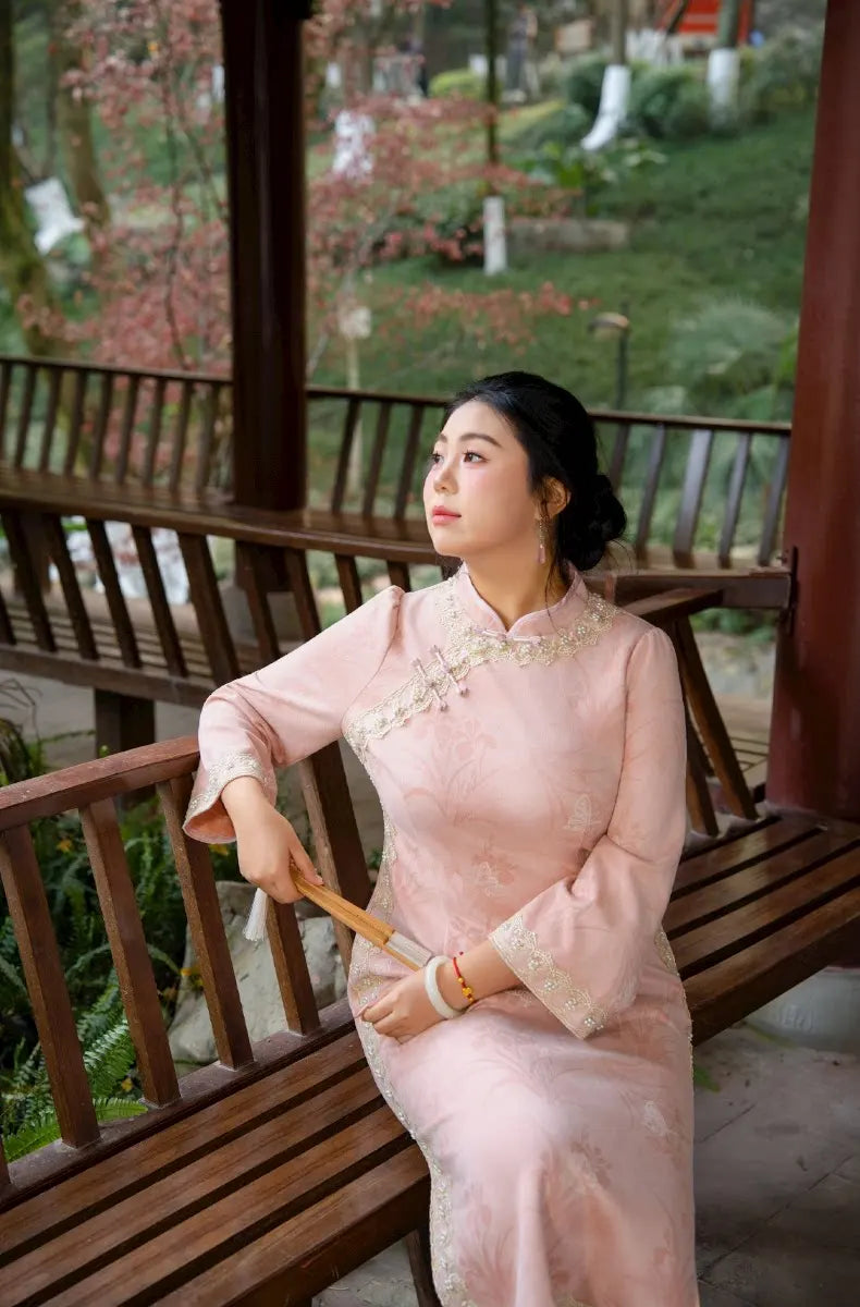 Woman in a pink traditional Plus Size Long Sleeve Pink Floral Qipao with Pearl Trim outfit sitting on a wooden bench with a garden view.
