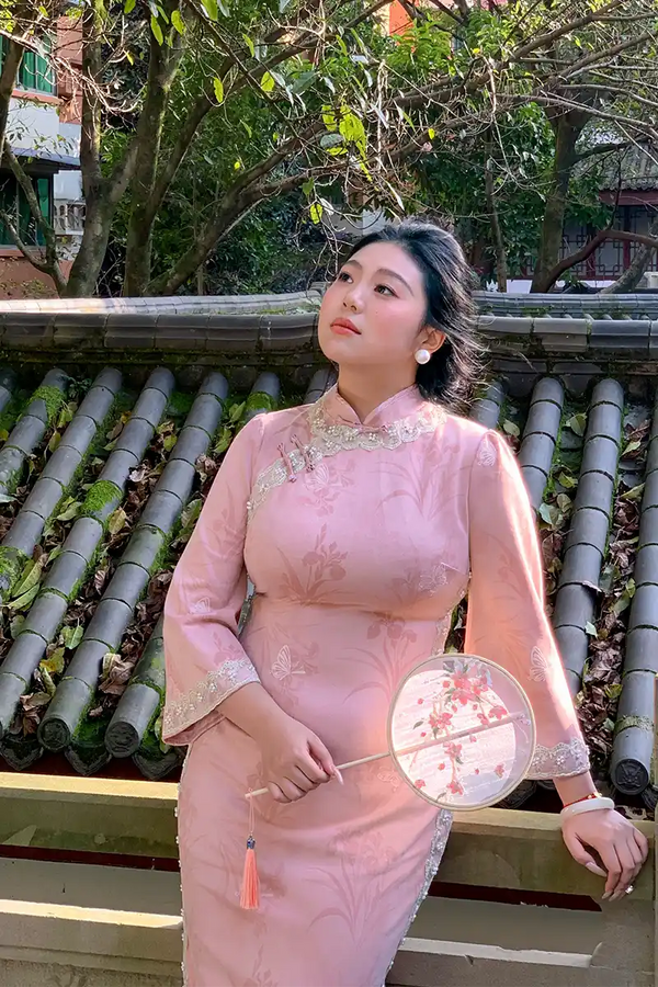 Woman in a pink traditional Plus Size Long Sleeve Pink Floral Qipao with Pearl Trim holding a fan, standing in front of a tiled roof and greenery.